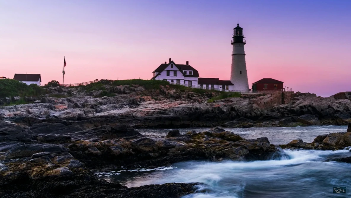 Lighthouse and keeper's house on rocky coastal outcrop at twilight with crashing waves below.