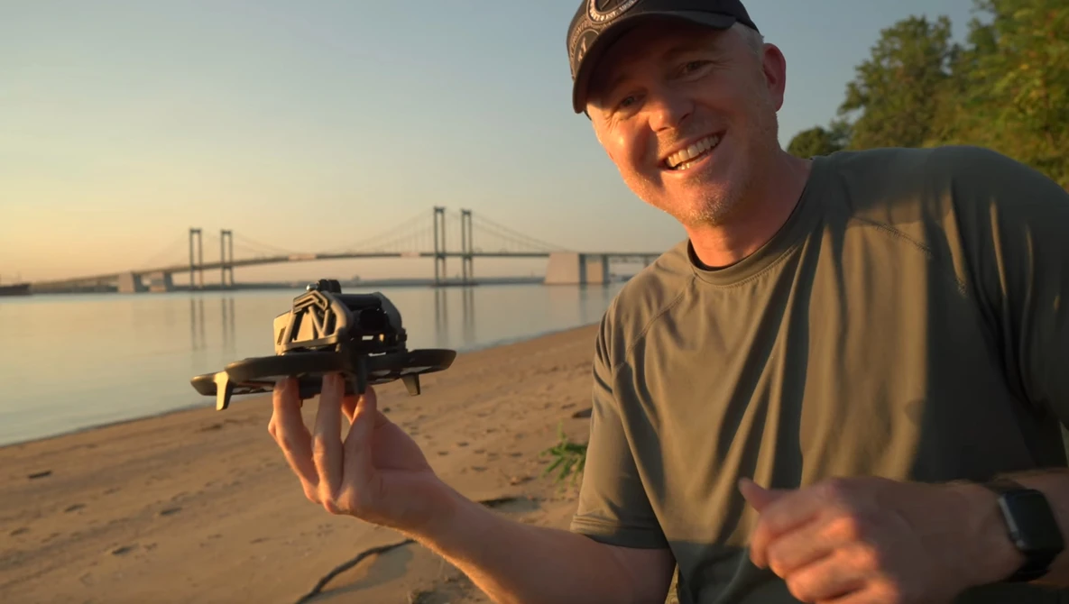 Smiling photographer holding a camera on a beach at golden hour with a suspension bridge visible across the water.