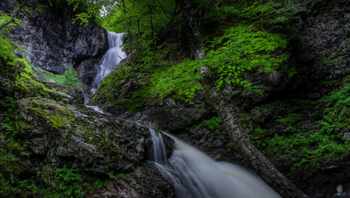 Multi-tiered waterfall cascading through moss-covered rocks in a densely forested ravine.