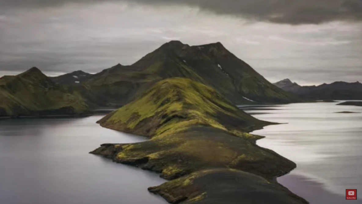 Moss-covered volcanic peaks rising from still water under overcast sky in Iceland.