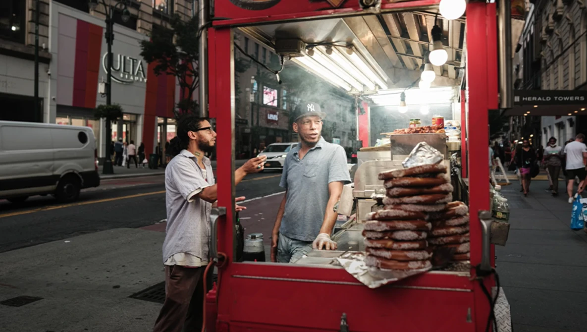 Street food vendor standing inside a red food cart on an urban street, with stacked packages of food visible and a customer approaching.