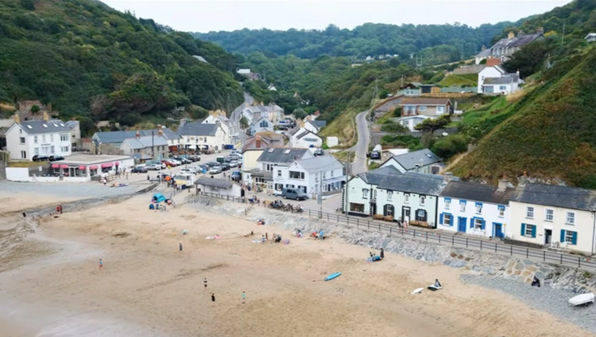 Aerial view of a crowded beach town nestled in green hills with Victorian-era buildings and a sandy shore.