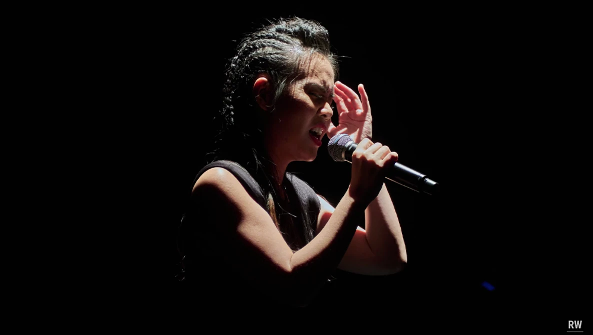 Female vocalist performing with microphone against dark background, wearing black sleeveless top.