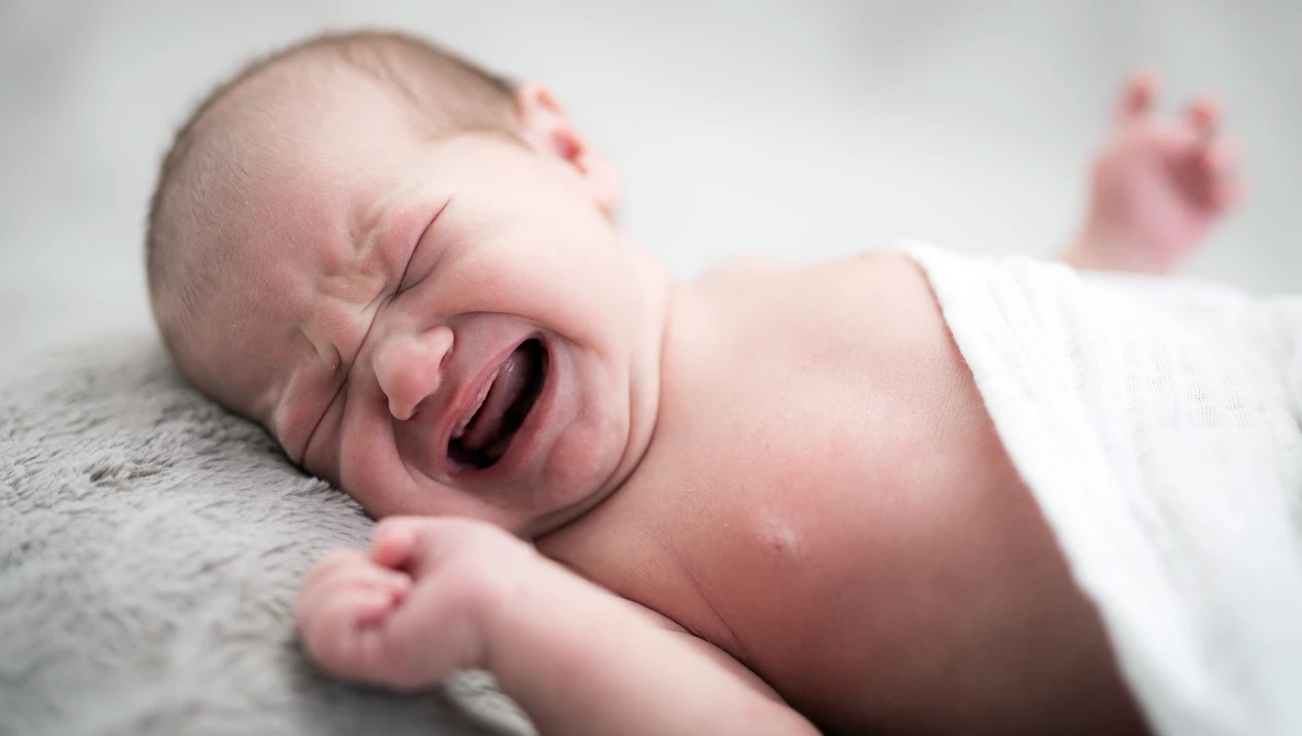 Newborn baby crying while lying on gray fabric with eyes squeezed shut.