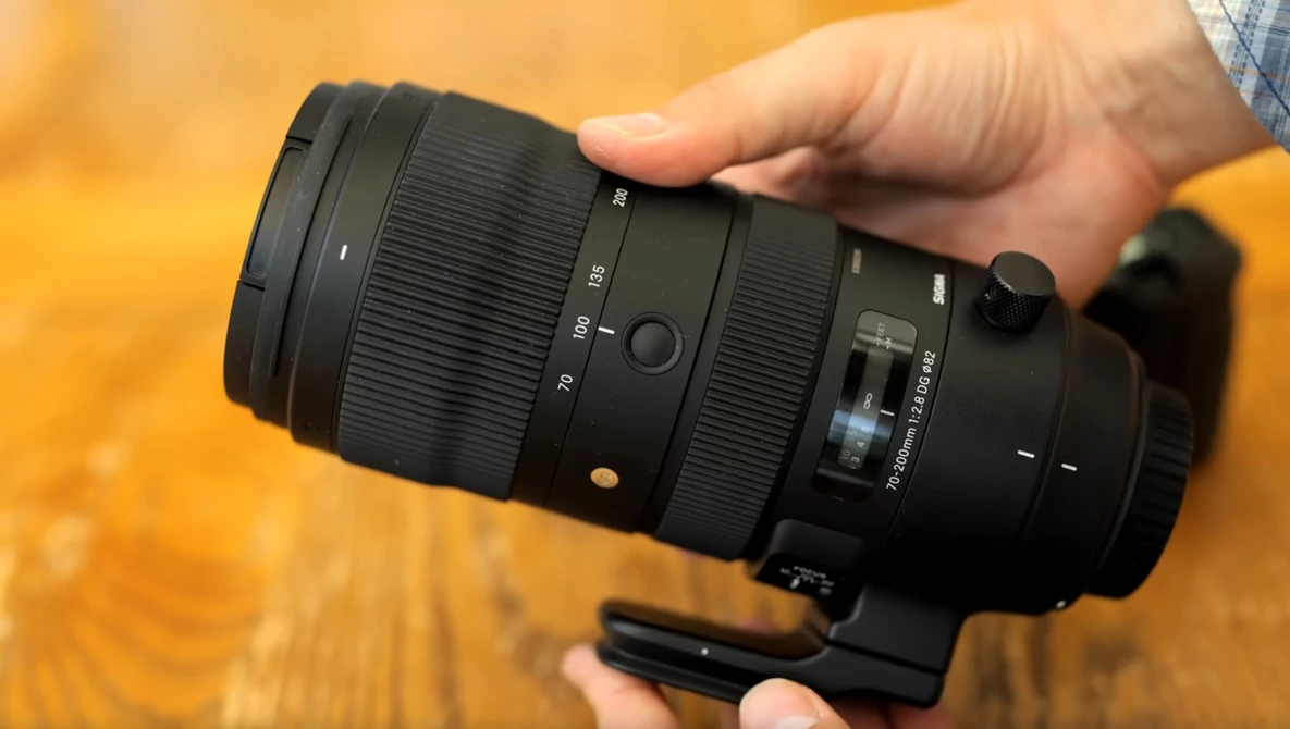 Hands holding a black telephoto lens with visible focal length markings and focus ring against a warm wooden background.