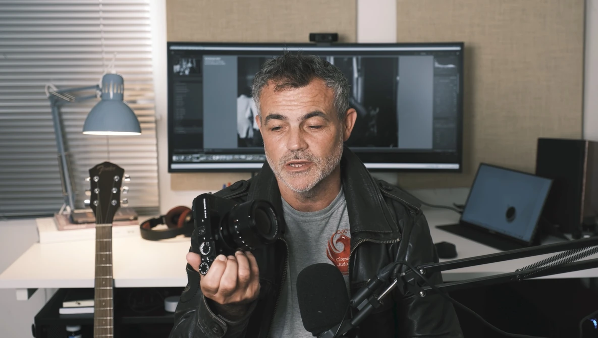 Man in black leather jacket speaking into microphone in home studio setup.