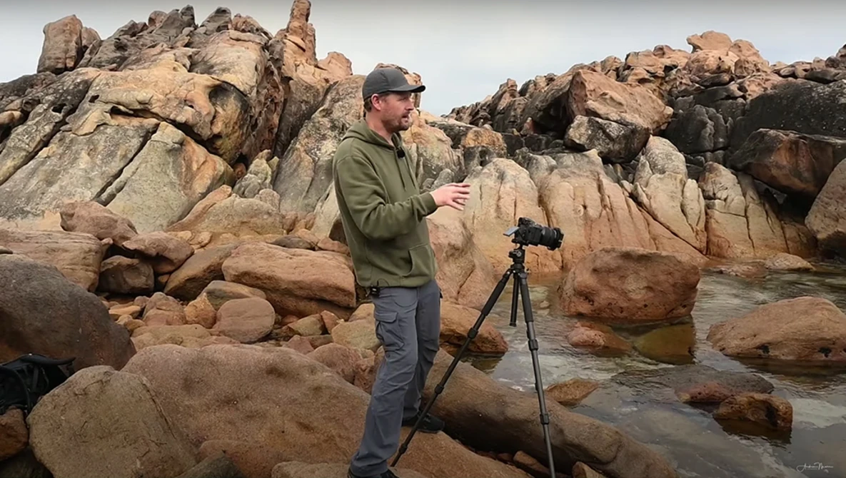 Photographer with camera on tripod standing among large granite boulders in rocky coastal landscape.
