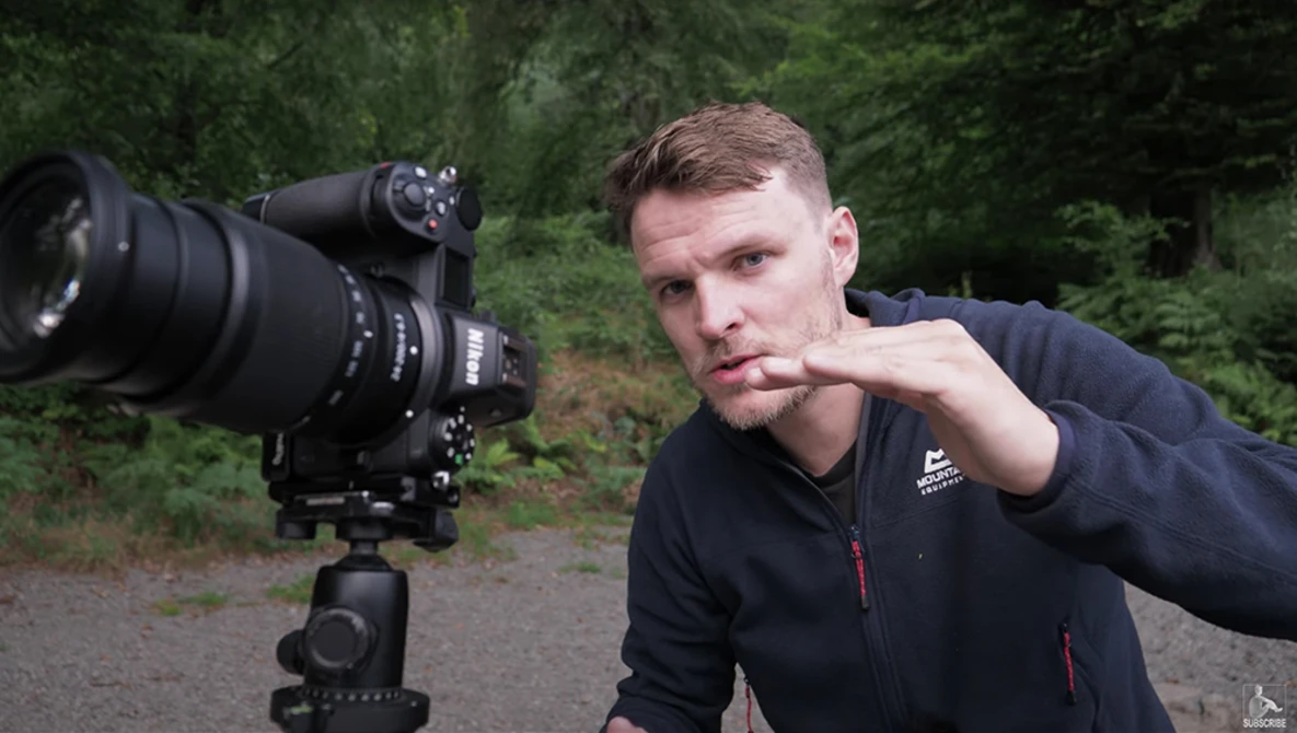 Male photographer posing thoughtfully beside a tripod-mounted camera with telephoto lens in a forested setting.