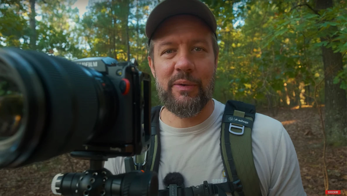 Videographer holding cinema camera with telephoto lens in forest setting during golden hour.