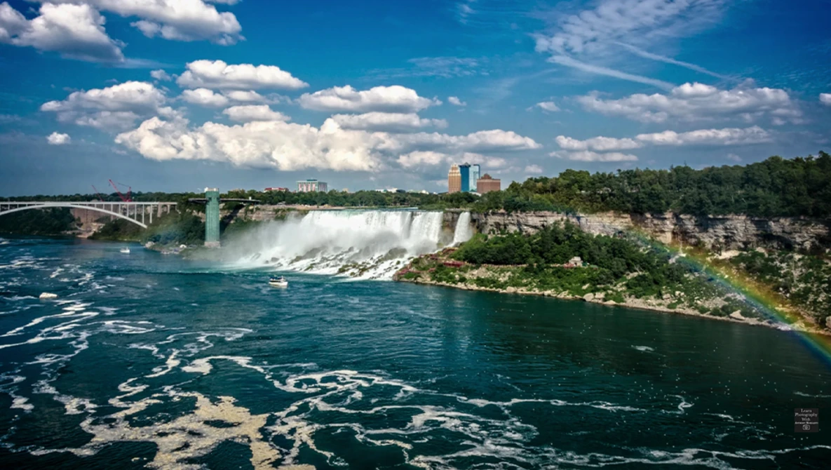 Massive waterfall cascading into turquoise river with bridge and buildings visible in background under blue sky.