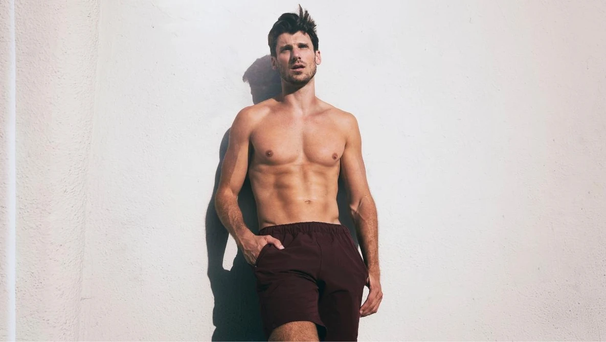 Shirtless male model with defined abdominal muscles posing against a white wall in studio lighting.