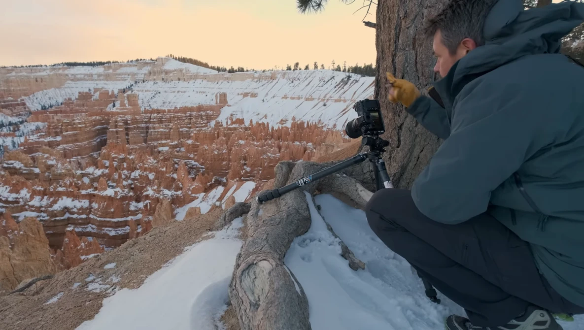 Photographer using a telephoto lens on a tripod at the edge of a snowy cliff overlooking a vast canyon landscape.