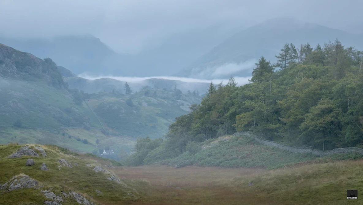 Misty valley landscape with forested hillside, distant mountains, and a small white building nestled in the valley below.