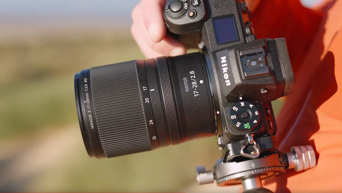 Mirrorless camera with telephoto lens mounted on red gimbal head, held against blurred landscape background.