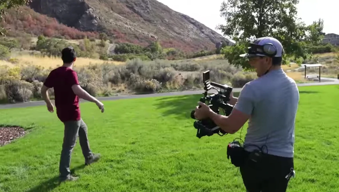 Cameraman operating a handheld cinema camera while filming a person in an open field with mountains and trees in the background.