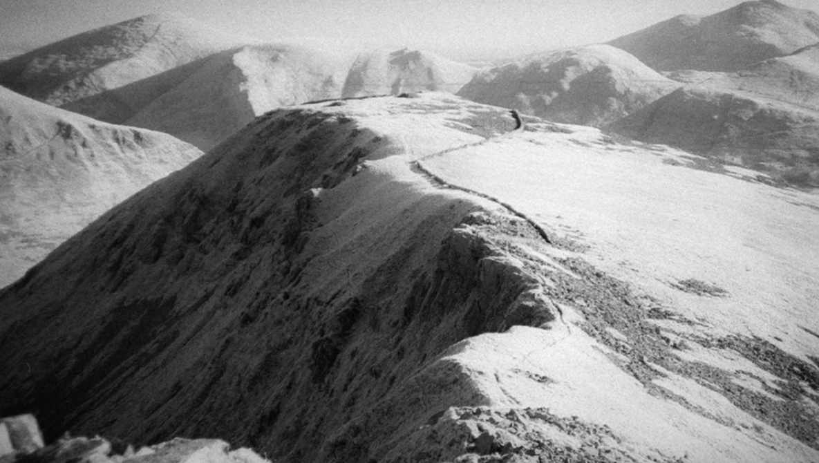 Snow-covered mountain ridge with exposed dark rock face under overcast alpine sky.