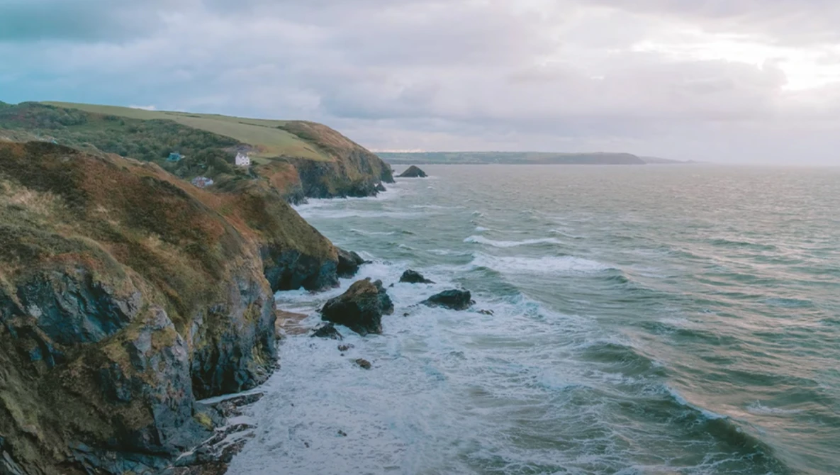 Dramatic coastal cliffs with rocky outcrops and crashing waves under overcast sky.