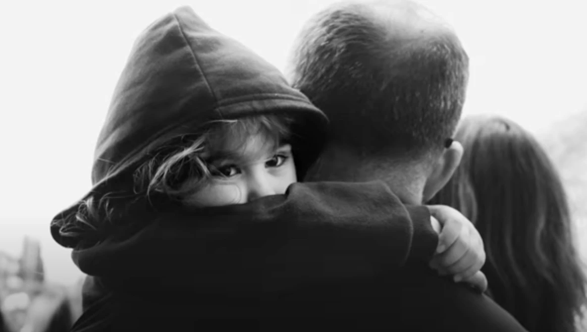 Child wearing a dark hoodie hugging an adult from behind in black and white.
