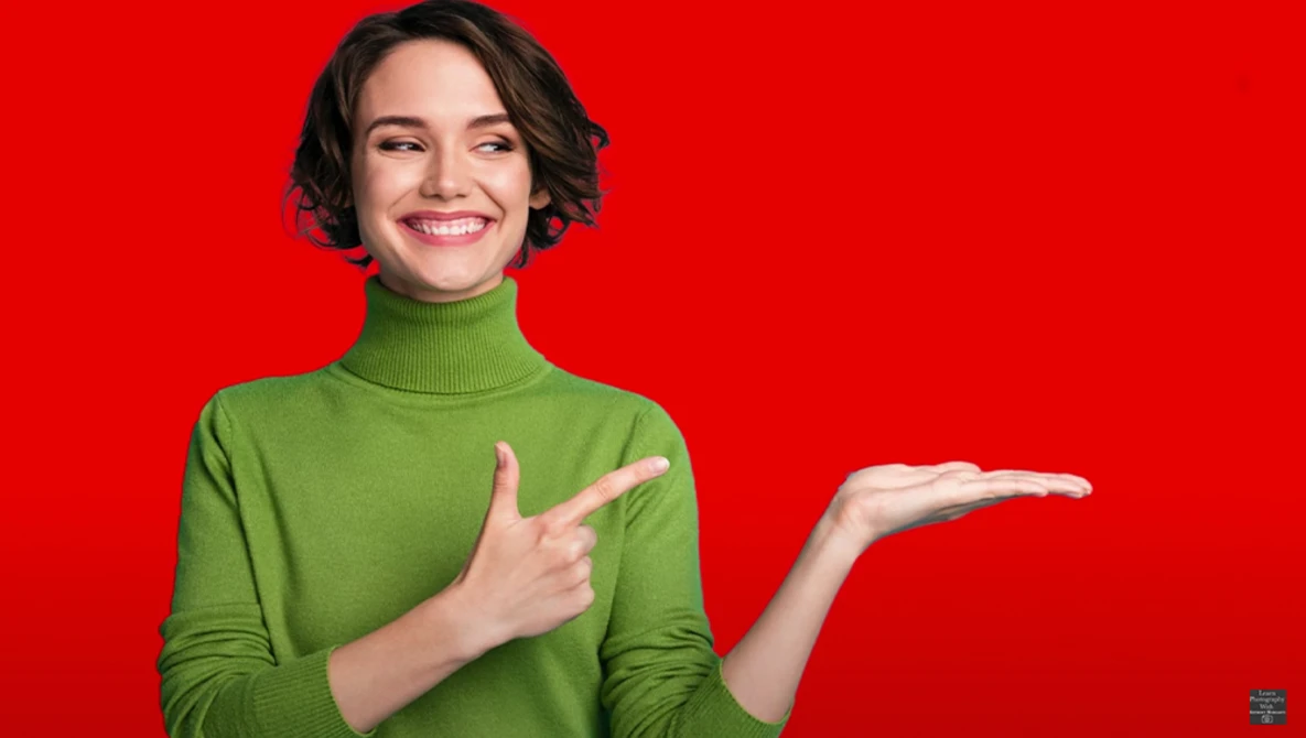 Woman in green turtleneck smiling against red background while pointing right.