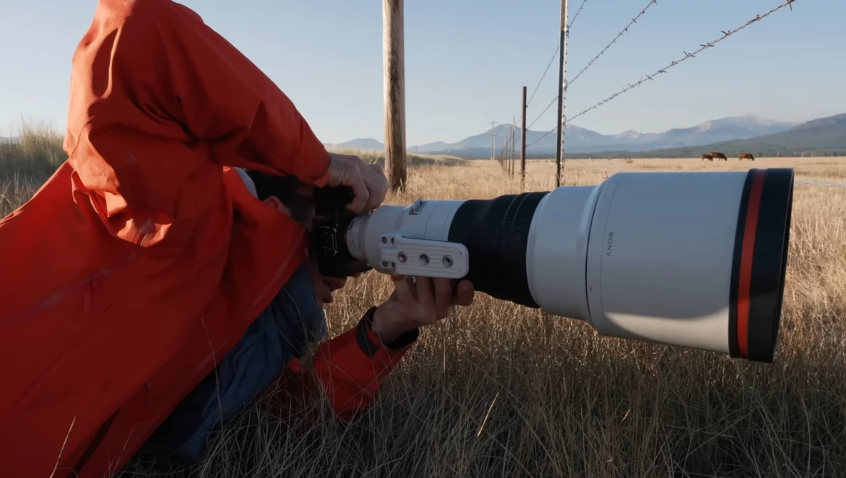 Photographer in red jacket using telephoto lens in open field with mountains in background.