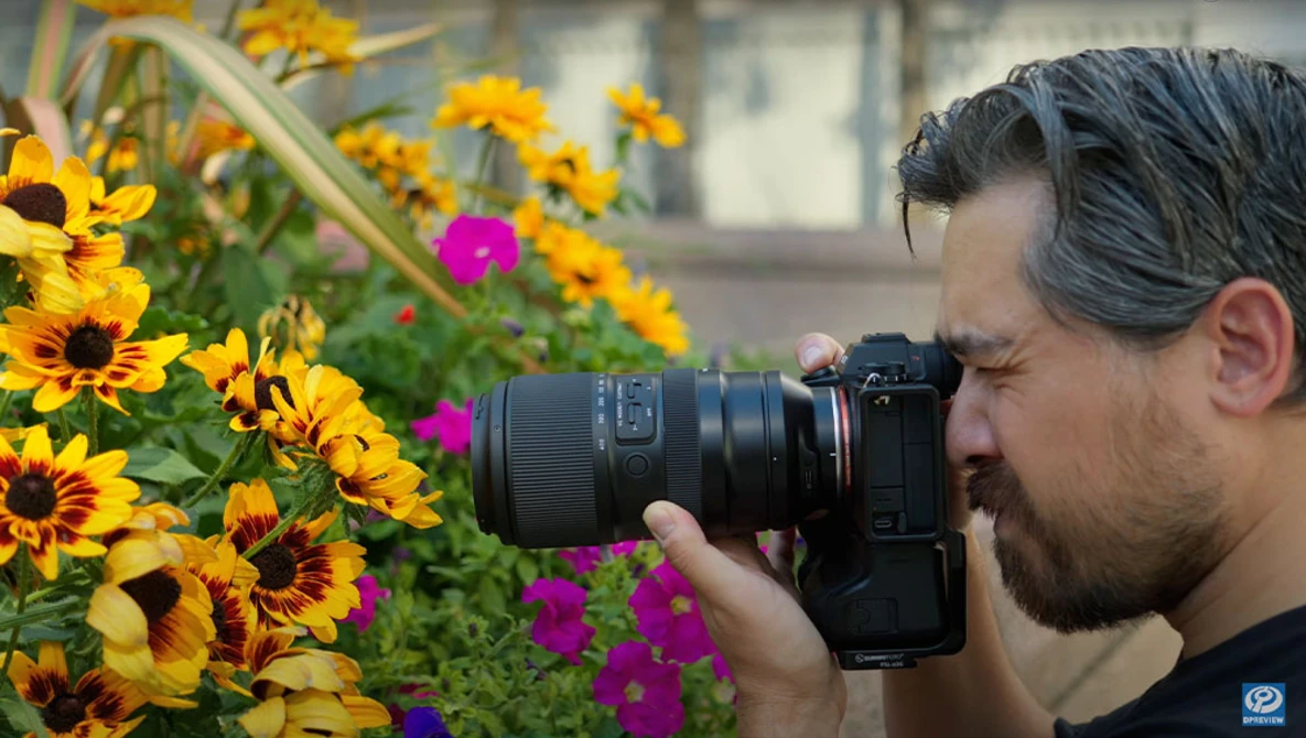 Photographer with mirrorless camera capturing close-up shots of yellow and magenta flowers in a garden setting.