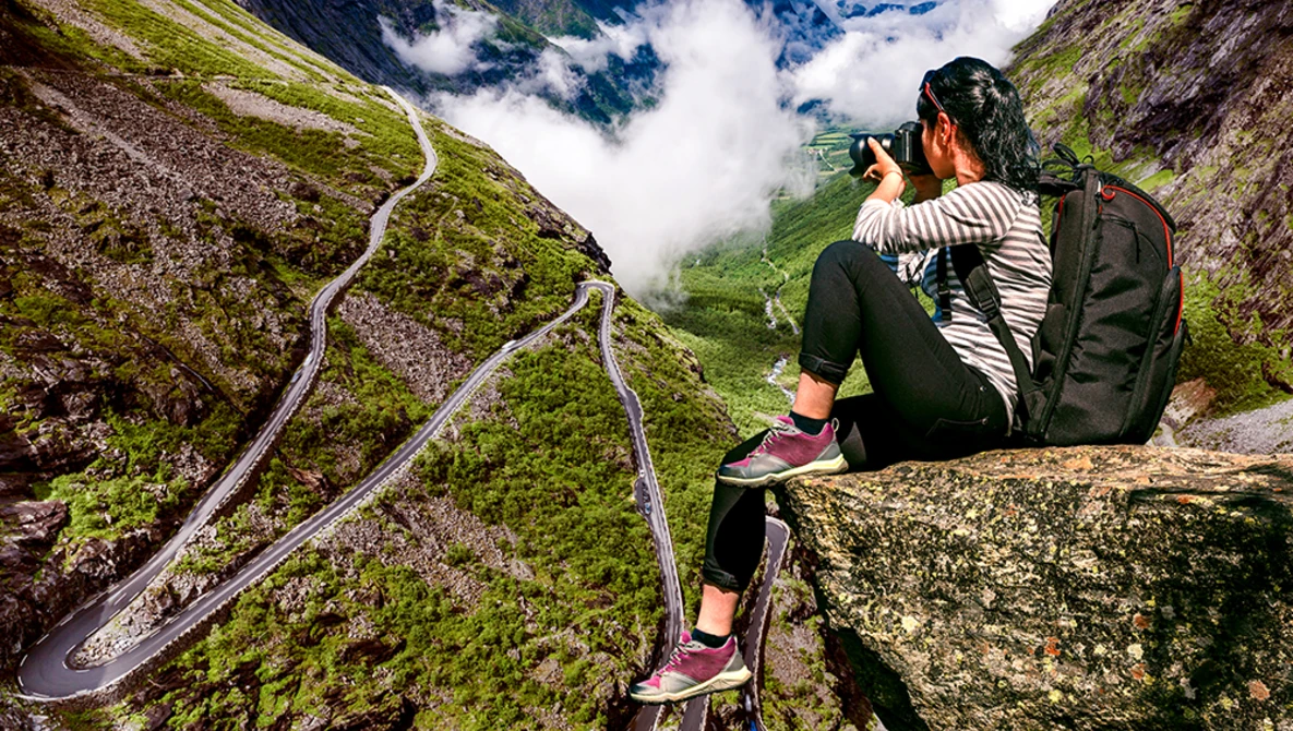Photographer on rocky outcrop capturing winding mountain road through valley below with camera.