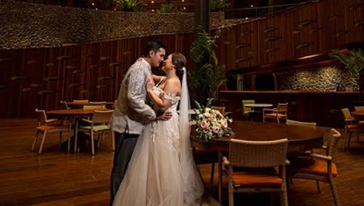 Bride and groom kiss in a modern wooden-walled venue with ambient lighting and floral arrangements.