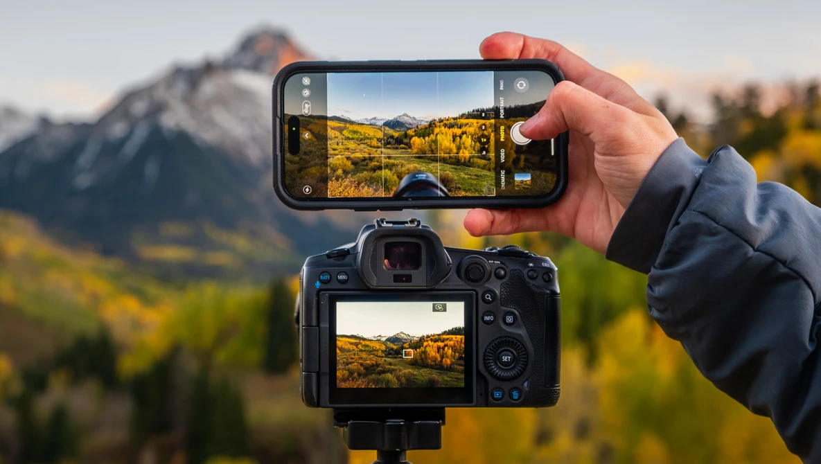 Smartphone and digital camera displaying identical mountain landscape scenes with golden vegetation and snow-capped peaks.