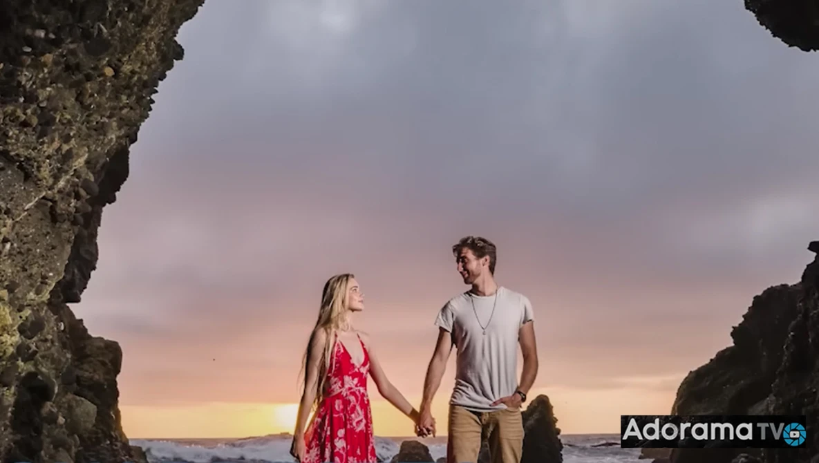 Couple holding hands on a beach at sunset, framed by large rock formations on either side.