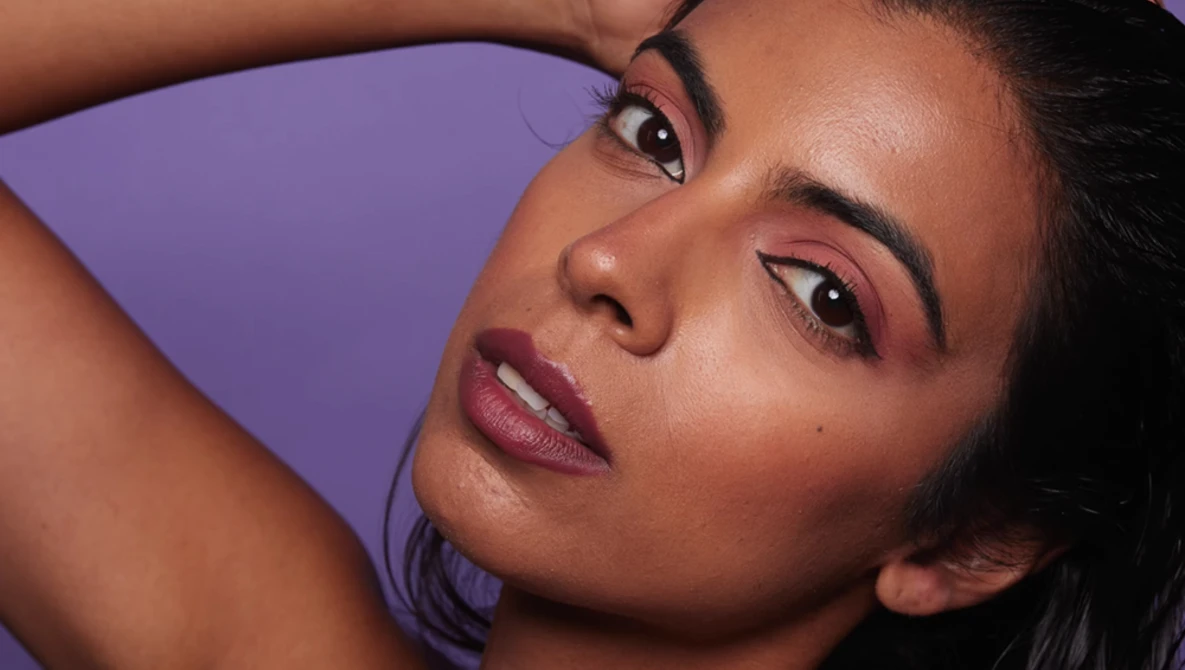 Woman with hand raised to head against purple background, wearing pink-toned makeup.