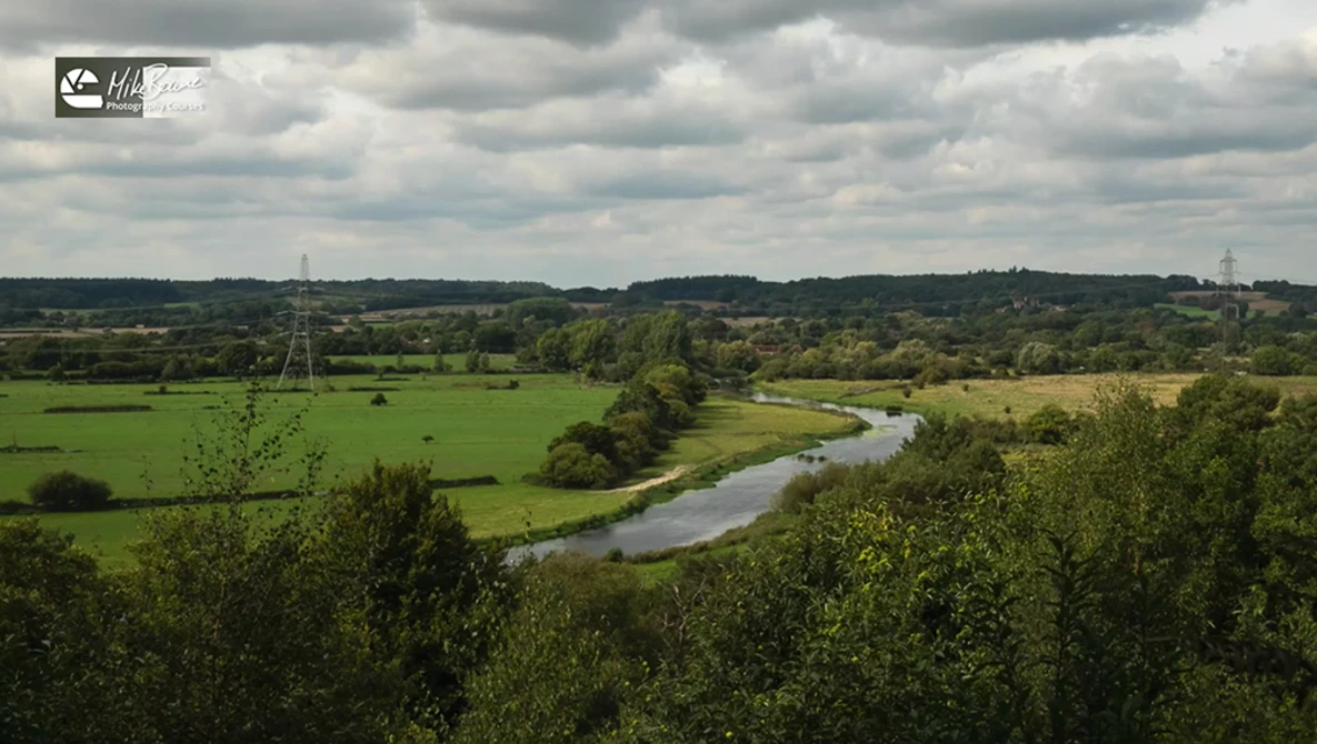 Meandering river cutting through pastoral English countryside with rolling hills, green fields, and distant woodlands under partly cloudy sky.