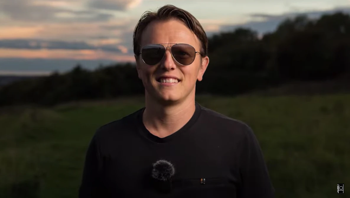 Man wearing sunglasses and dark t-shirt smiling outdoors during golden hour.