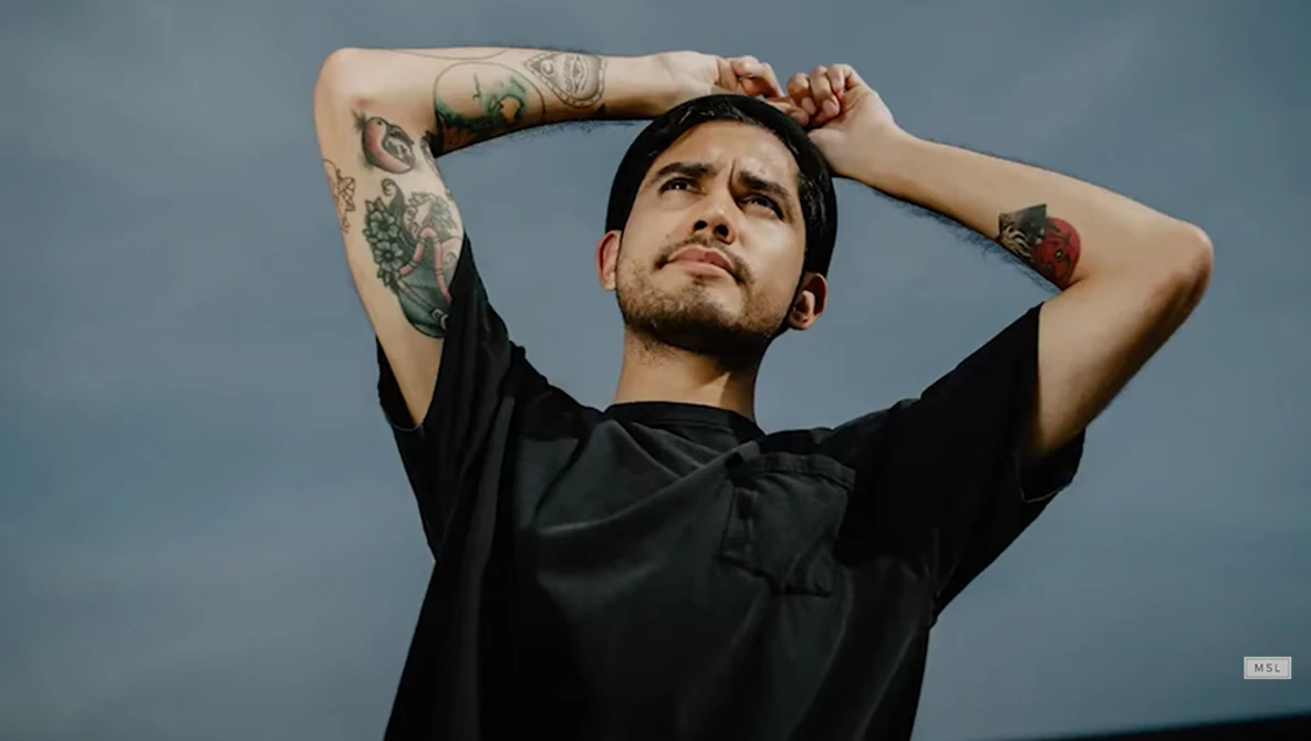 Tattooed man in black shirt with hands behind head against blue-gray sky background.
