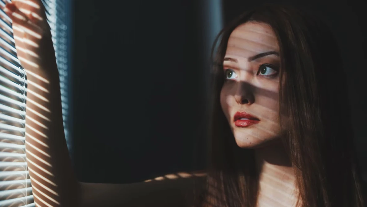 Woman with long dark hair looking toward a window with venetian blinds casting striped shadows across her face.