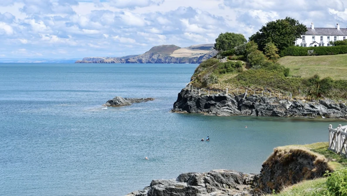 Turquoise coastal cove with rocky outcrops, white house on green headland, and distant cliffs across blue water.