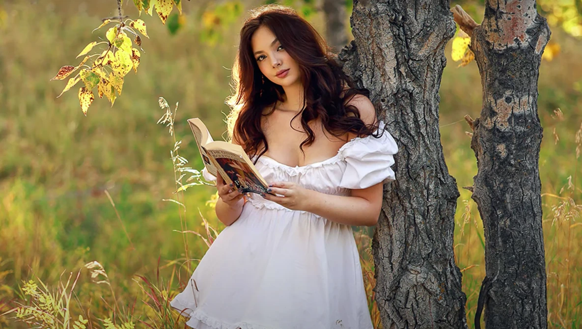 Woman in white off-shoulder dress holding dried corn stalks beside a weathered tree trunk.