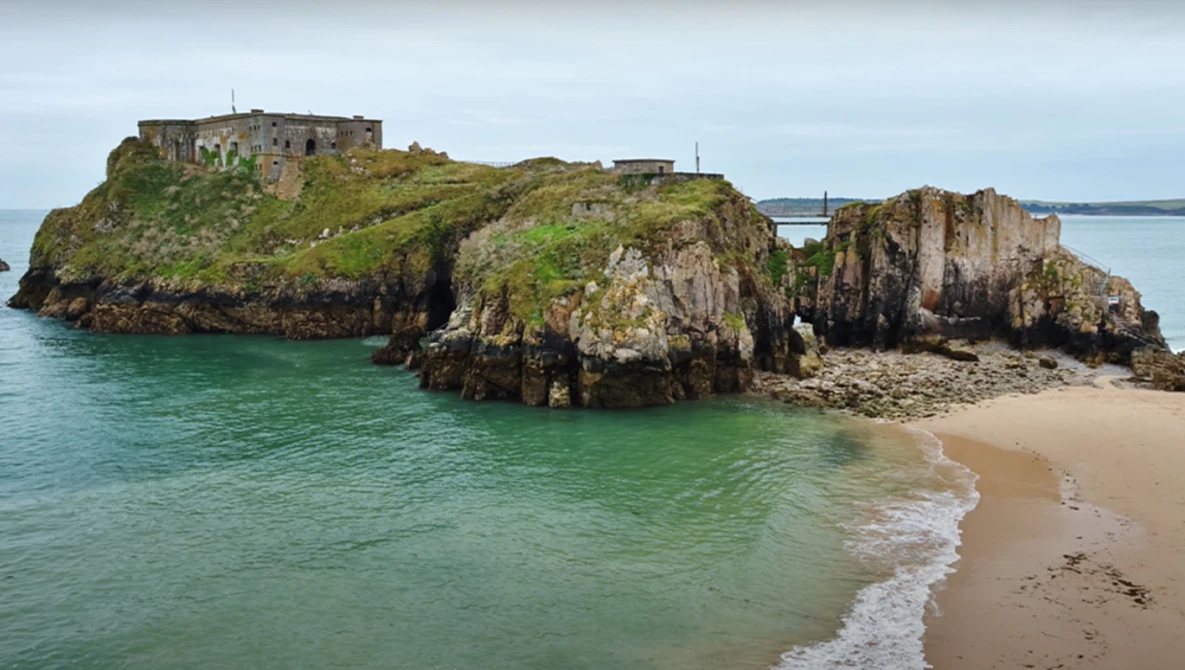 Coastal fortress ruins on a rocky island connected by a causeway, with turquoise water and sandy beach.