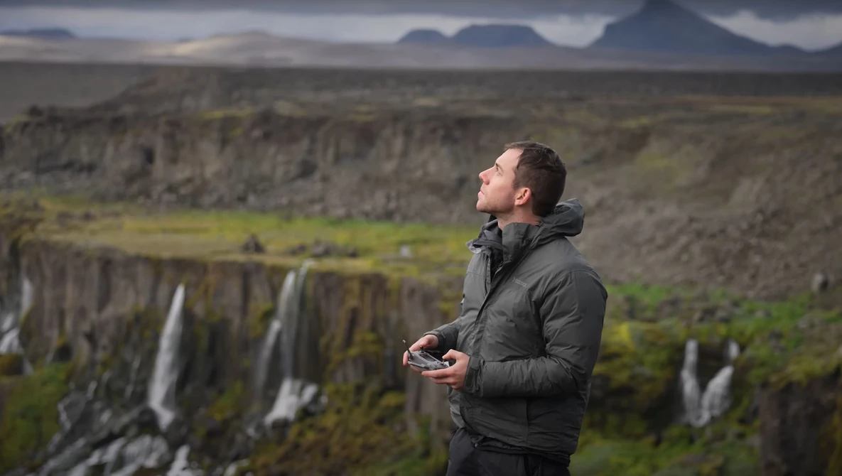 Man holding a drone controller overlooking a vast landscape with waterfalls and basalt cliffs.