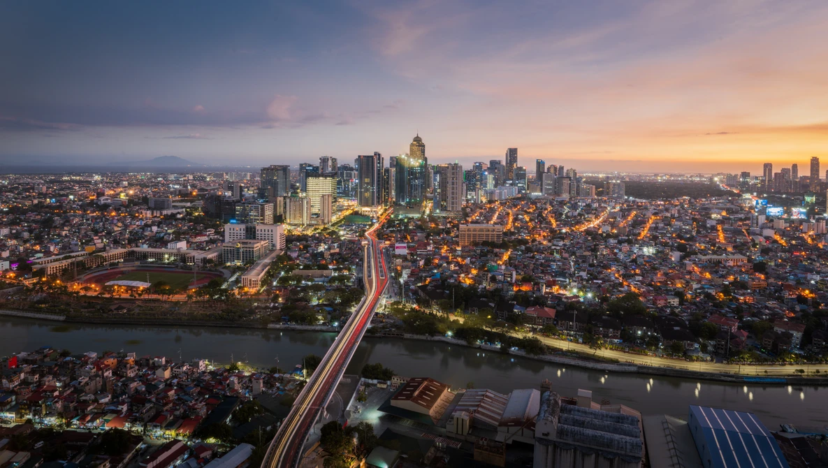 Cityscape at dusk with illuminated skyscrapers, a river, and light trails from traffic on an elevated highway.