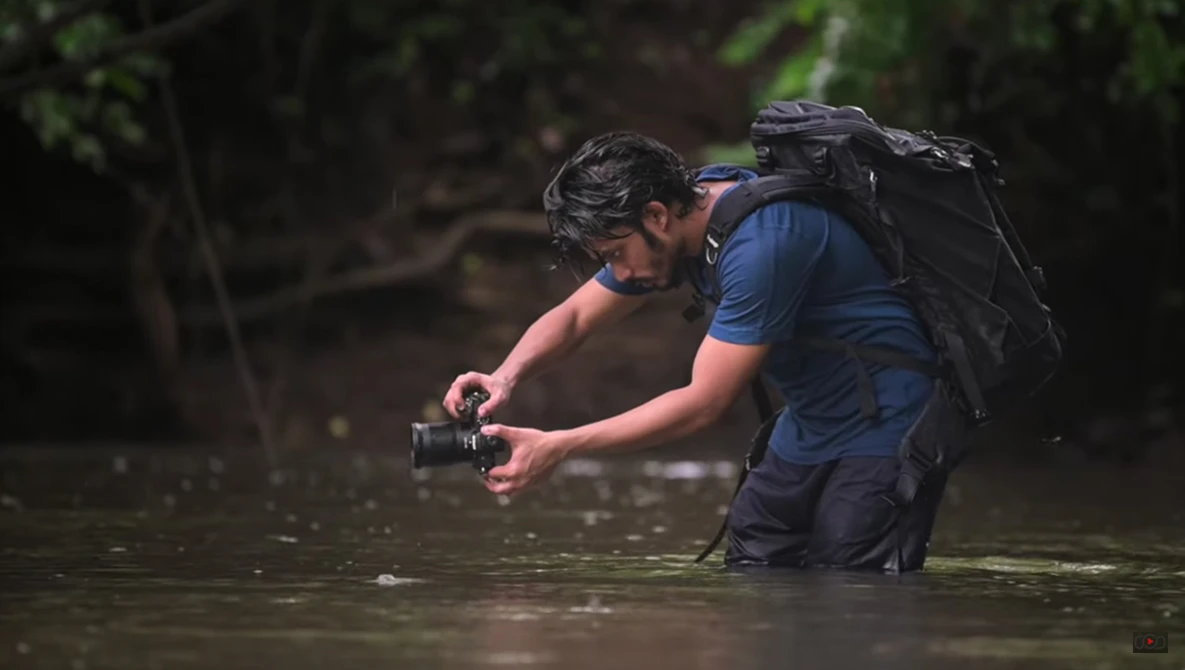 Photographer wading in shallow water while operating a camera, wearing a blue shirt and black backpack.