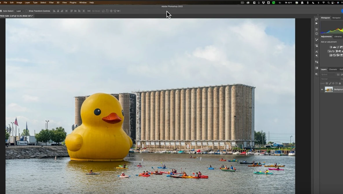 Giant yellow rubber duck floating on water with industrial grain elevator and kayakers in background.