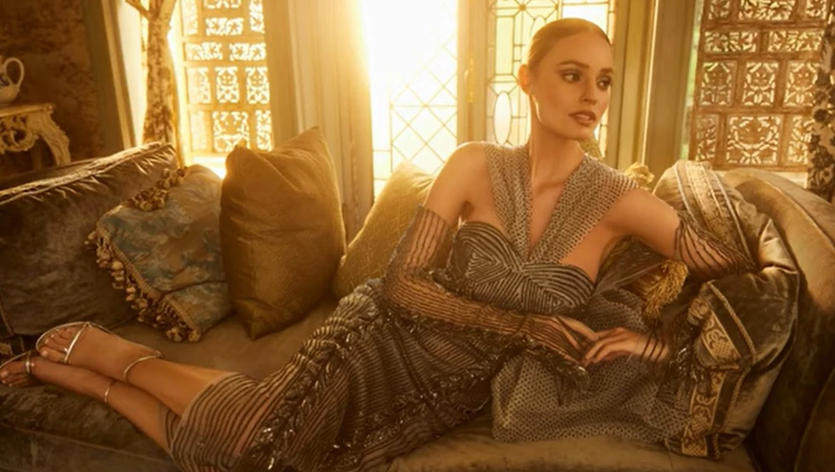 Woman in beaded wrap dress reclining on ornate sofa with decorative pillows and bright window light behind her.