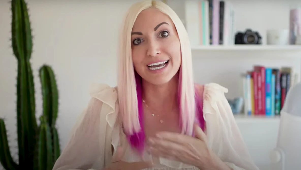 Woman with platinum blonde and pink ombre hair speaking to camera in bright, modern interior space.