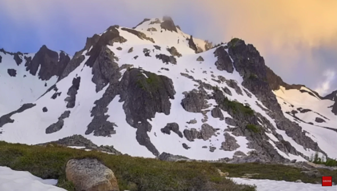 Snow-covered alpine peak with exposed dark rock faces and a lush green meadow in the foreground during golden hour.