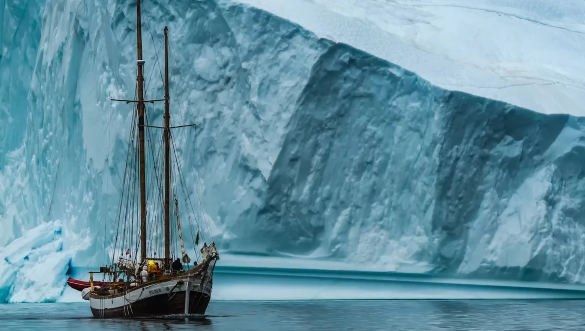 Wooden sailing vessel anchored in calm glacial waters beneath towering ice cliffs.