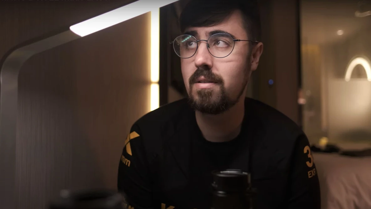 Man wearing glasses and dark clothing holds a camera in a modern interior with warm ambient lighting.