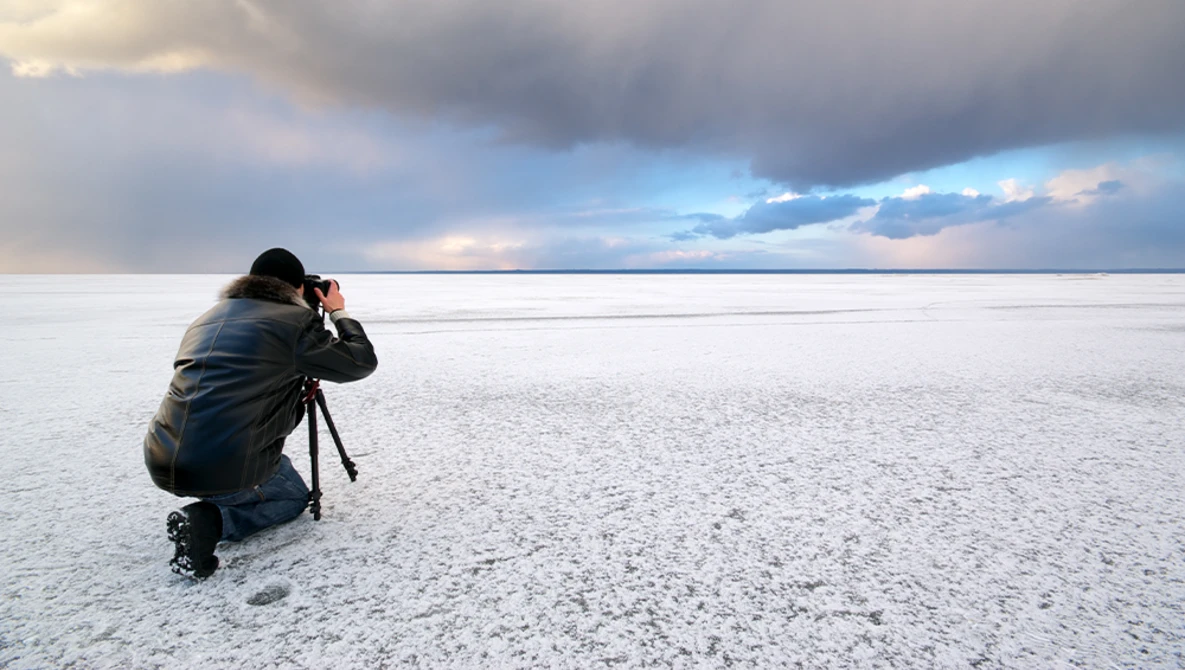 Photographer crouching on a salt flat with a camera on a tripod, framing a dramatic cloudy sky at sunset.