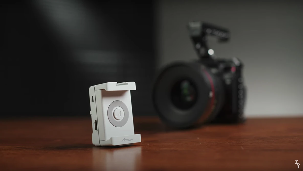 White compact camera device in sharp focus on wooden surface with professional camera equipment blurred in background.