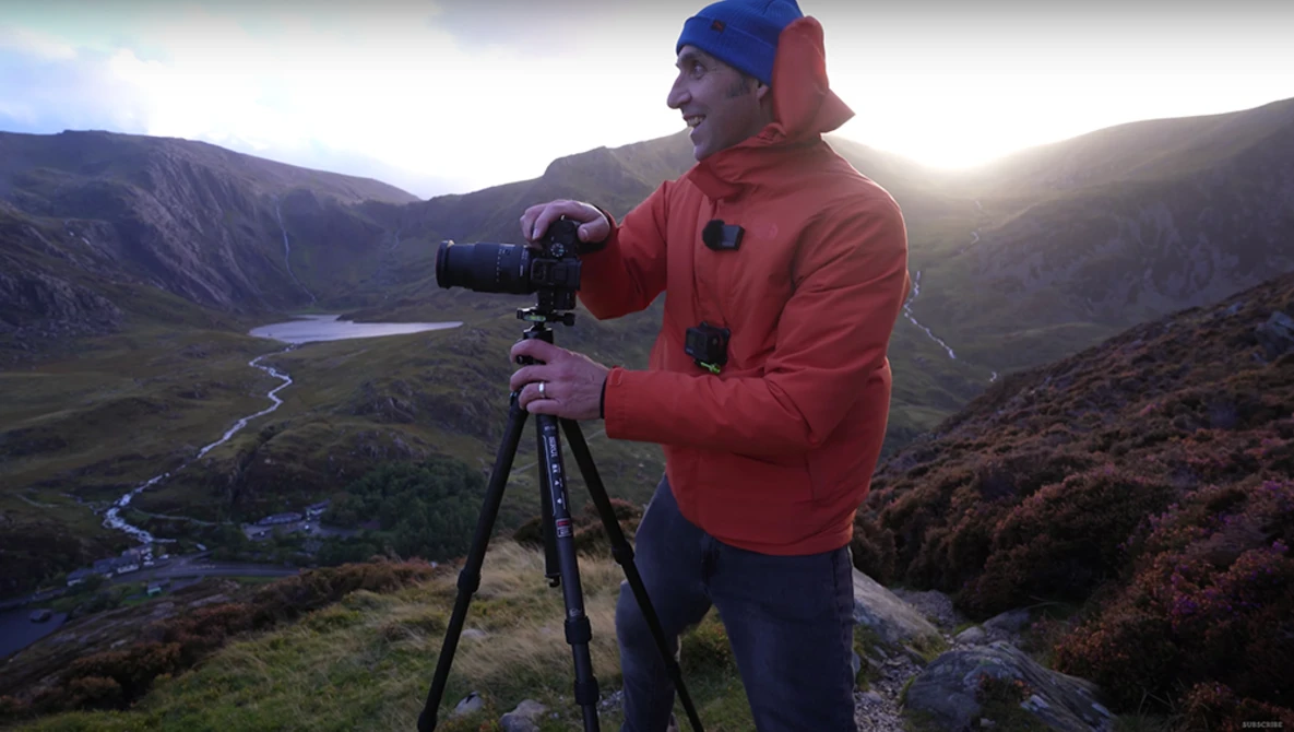 Photographer in red jacket and blue beanie operating a camera on tripod in mountainous valley landscape.