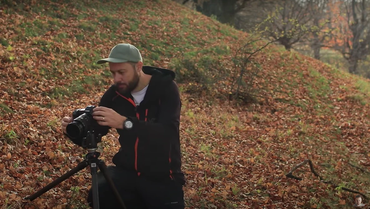 Bearded photographer adjusting a cinema camera mounted on a tripod in an autumn forest setting.